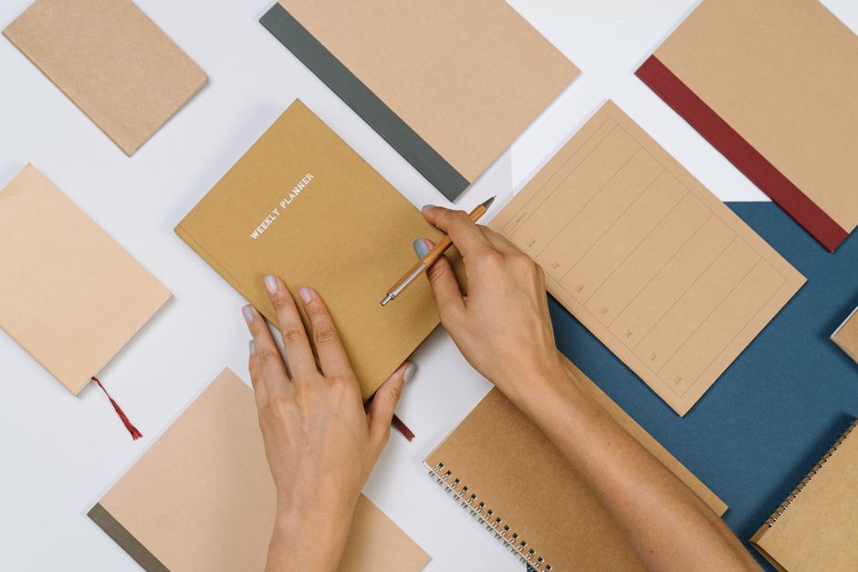 Hands holding a pen and a closed weekly planner among open notebooks